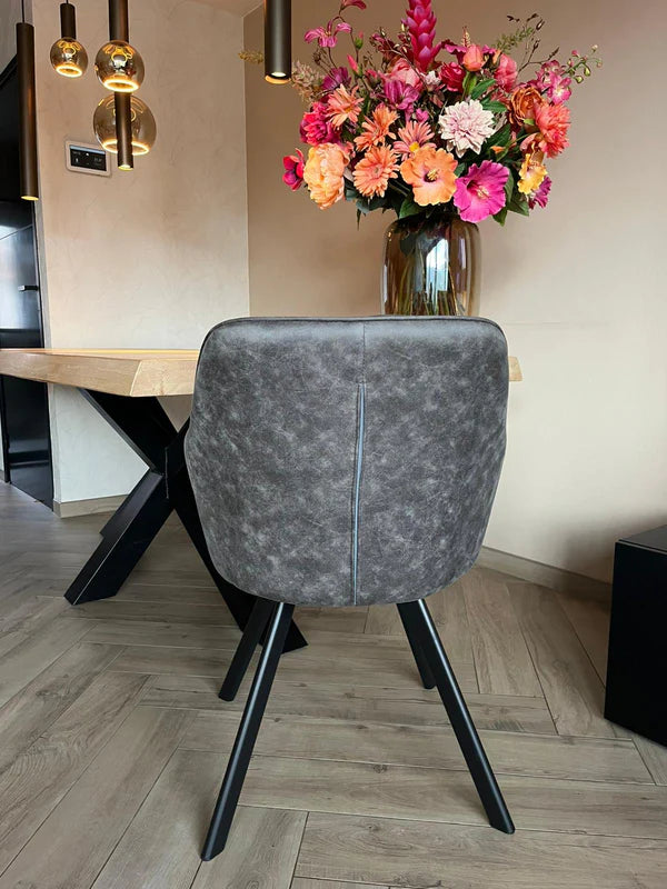 Eetkamerstoel Draaibaar 'Brione' met donkere stoffering en zwart metalen poten, naast een houten tafel met bloemenvaas en moderne hanglampen.