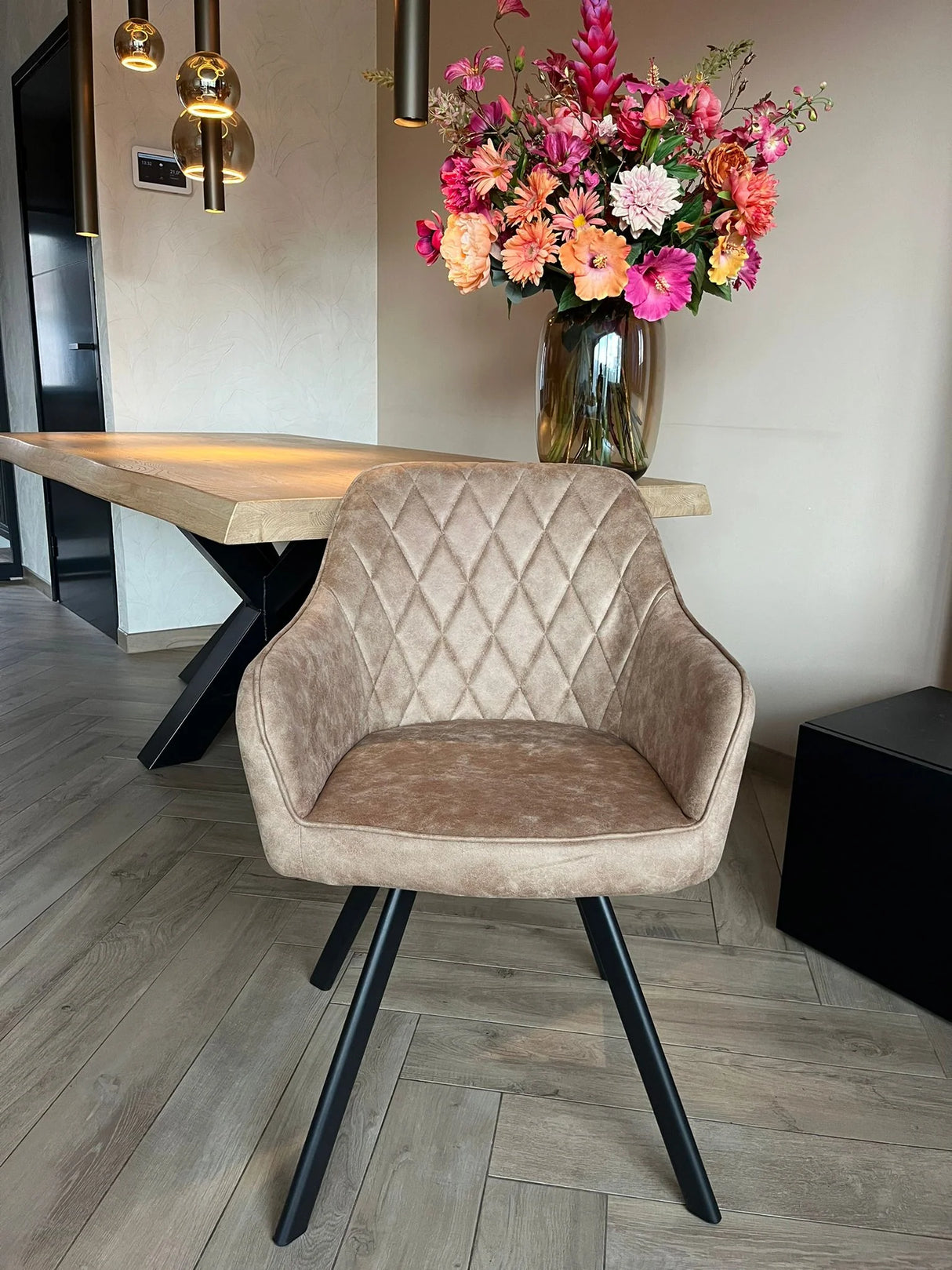 Eetkamerstoel Draaibaar 'Brione' met beige microvezel stof en zwart metalen poten, naast een houten tafel en bloemenvaas, verlicht door hanglampen.
