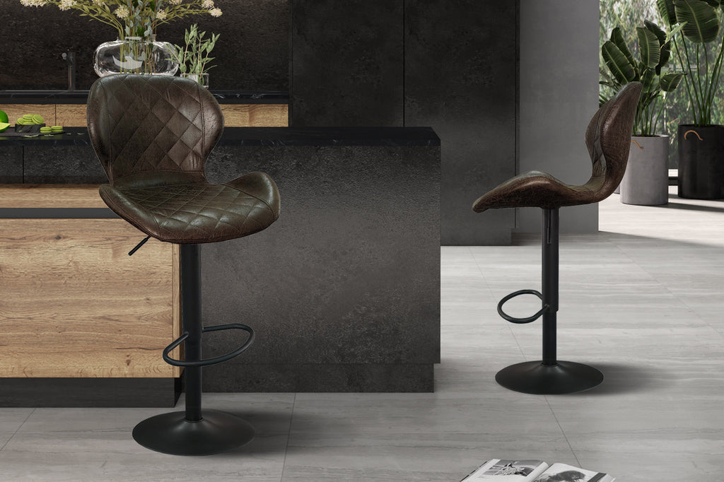 Twee bruine barkrukken 'Malaga' met PU-leer en metalen basis, geplaatst in een kamer met een vaas en plant als decor.