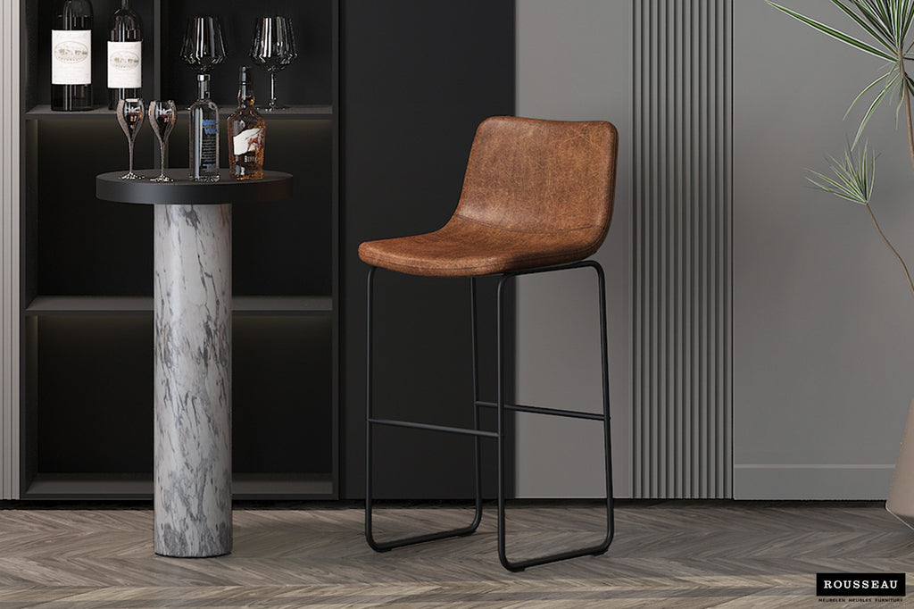 Barkruk 'Mancia' PU Cognac naast een tafel, met bruine zitting en zwarte poten, perfect voor een stijlvolle interieurinrichting. Verkrijgbaar bij Furndaily.