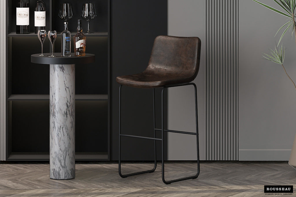 Barkruk 'Mancia' PU Donkerbruin naast een tafel, met een zwart metalen frame, perfect voor stijlvolle interieurs. Verkocht als set van twee.