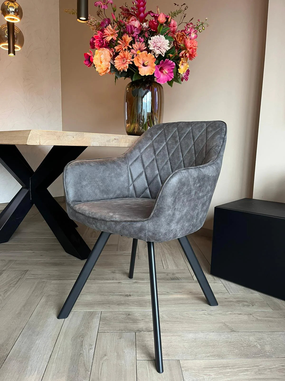 Eetkamerstoel Draaibaar 'Brione' in grijs microvezel met diamantstiksels, slank zwart metalen frame, naast een lichte eiken tafel met bloemenvaas.