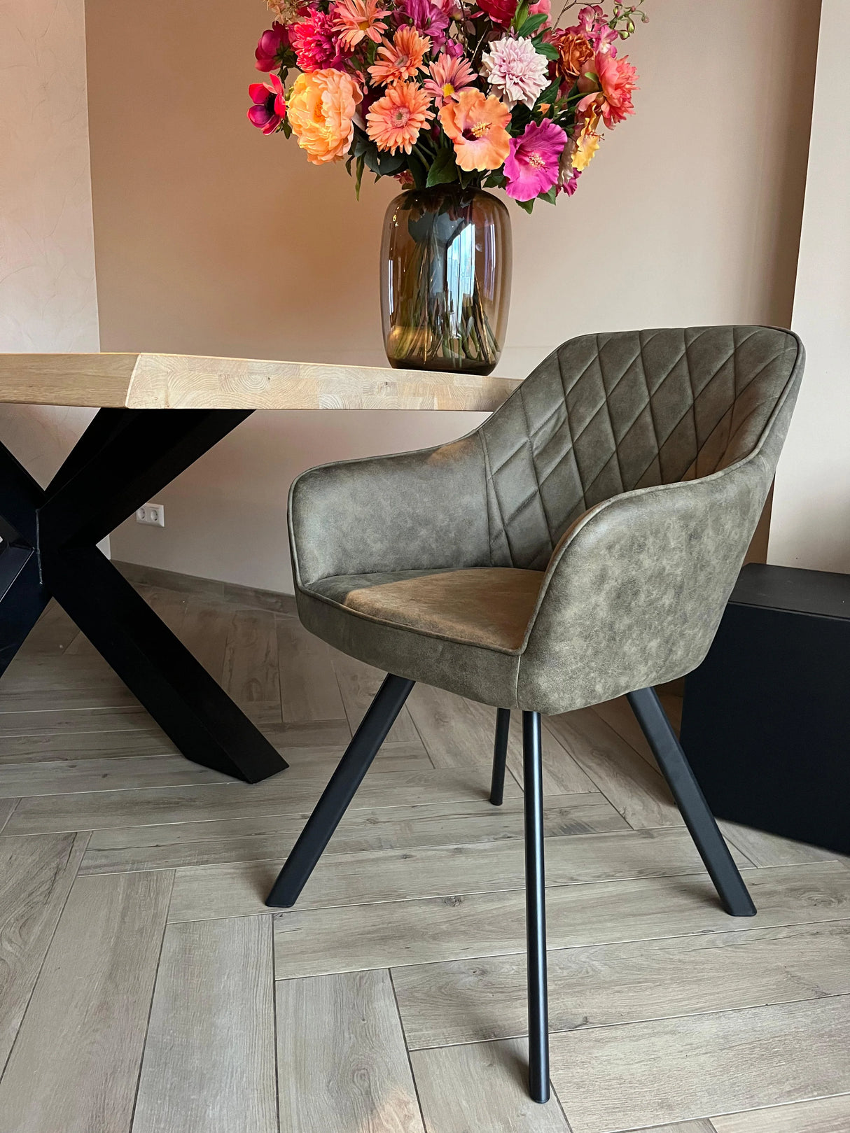 Eetkamerstoel Draaibaar 'Brione' in olijfgroen met diamantstiksel en zwarte metalen poten, naast een tafel met vaas en bloemen, op visgraatvloer.