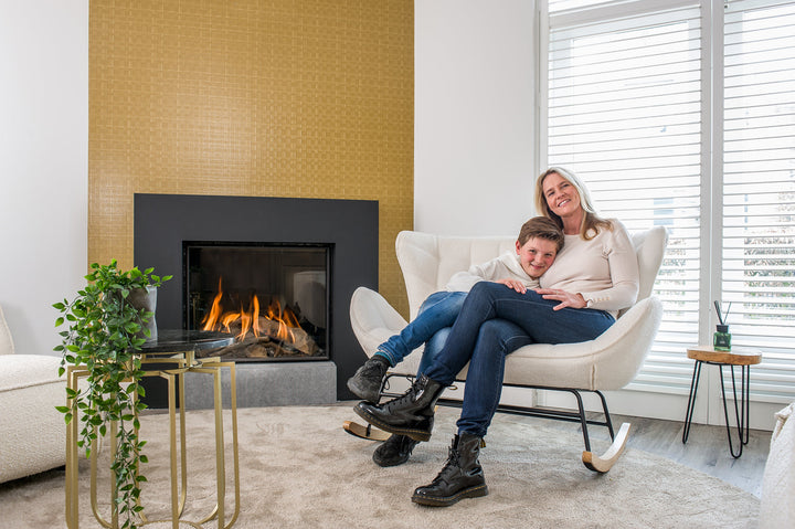 Vrouw en jongen zitten op de Aemely® Schommelbankje Duo, bekleed met witte schapenstof, voor een haard in een gezellige woonkameromgeving.